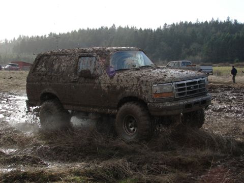 1996 Ford Bronco - This SUV is covered in mud while driving through a field with trees in the background.