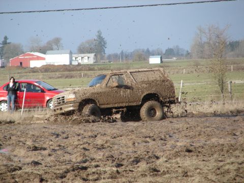1996 Ford Bronco - A mud-covered SUV struggles in a muddy field, with a red car and a bystander observing.