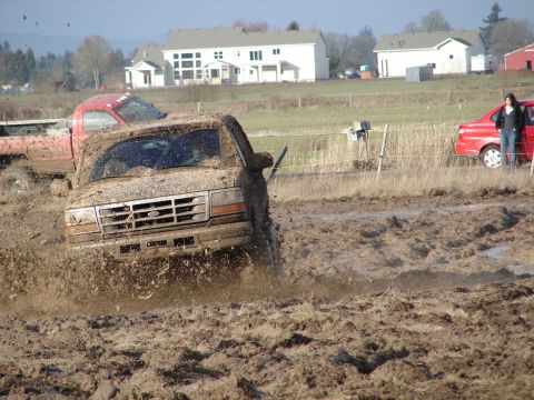 1996 Ford Bronco - A muddy Ford truck navigates a challenging off-road course, with other vehicles and houses in the background.