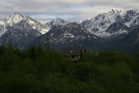 2003 Jeep TJ - A remote cabin sits amidst lush greenery, dwarfed by , snow-covered peaks under a cloudy sky.