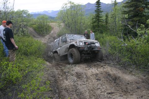 2003 Jeep TJ - A Suzuki Samurai tackles a challenging muddy incline during an off-road event, observed by several people.