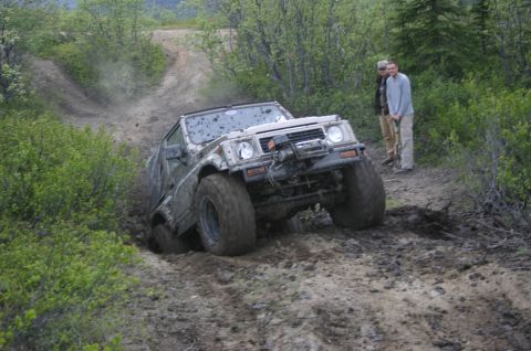 2003 Jeep TJ - Two men watch as a heavily soiled off-road vehicle struggles on a challenging dirt track.