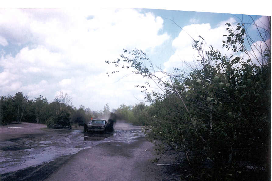 Dark SUV mudding through deep puddles on a muddy off-road trail lined with brush