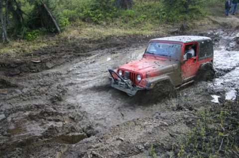 2003 Jeep TJ - A red 2003 Jeep TJ Wrangler navigates a challenging muddy trail, with its front end submerged in water and mud.
