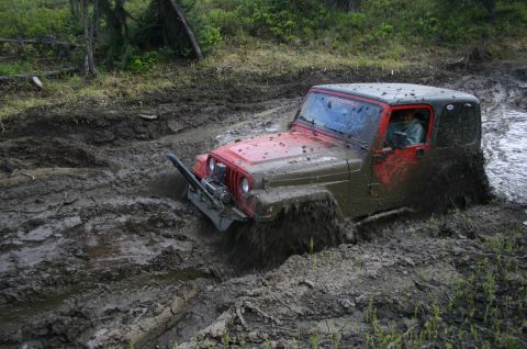 2003 Jeep TJ - A red Jeep TJ is struggling to get through a muddy off-road trail, with mud splashing around its tires.