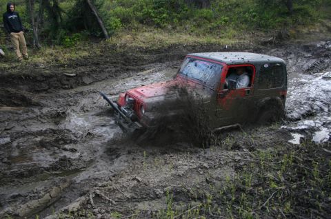 2003 Jeep TJ - A red Jeep Wrangler TJ navigates a challenging muddy trail, with mud splashing around it.