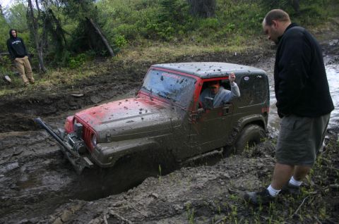 2003 Jeep TJ - A red 2003 Jeep TJ is stuck deep in mud during an off-road event, with a driver waving from the window.