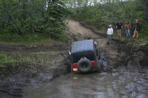 2003 Jeep TJ - A Jeep TJ navigates a challenging muddy water crossing on an off-road trail, with onlookers observing the action.