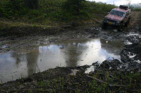 2003 Jeep TJ - A red Jeep Cherokee XJ tackles a challenging muddy obstacle course during an off-road event.