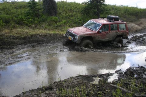 2003 Jeep TJ - A red Jeep Cherokee navigates a muddy trail, with its tires deep in a water-filled rut.