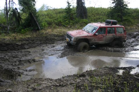 2003 Jeep TJ - A red Jeep Cherokee XJ navigates a challenging muddy terrain, with one wheel deep in a water-filled rut.