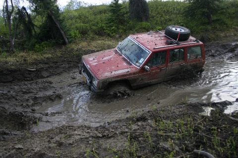 2003 Jeep TJ - A red Jeep Cherokee XJ navigates a muddy trail, getting stuck in deep mud.