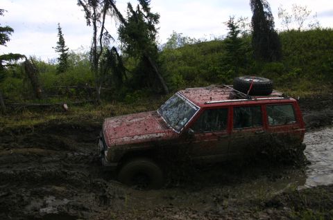 2003 Jeep TJ - A red Jeep Cherokee XJ is deeply stuck in mud, with its tires submerged and mud splashing around it.