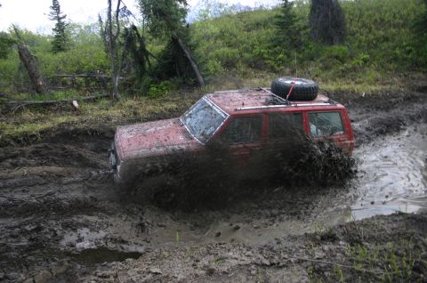 2003 Jeep TJ - A red Jeep Cherokee XJ navigates a muddy trail, kicking up a large spray of mud.