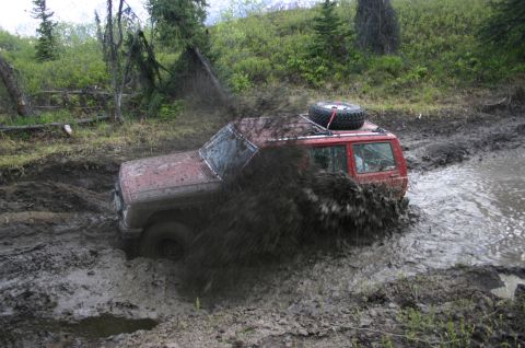 2003 Jeep TJ - A red 2003 Jeep TJ Wrangler navigates a muddy trail, kicking up a large spray.