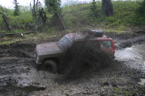 2003 Jeep TJ - This red SUV is deeply stuck in a muddy trail, with a large amount of mud being displaced.