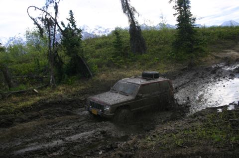 2003 Jeep TJ - A red Jeep Cherokee XJ navigates a muddy trail, with snow-capped mountains visible in the distance.