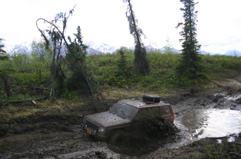 2003 Jeep TJ - A Jeep Cherokee is stuck deep in the mud on an off-road trail, with water splashing around it.
