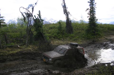 2003 Jeep TJ - A maroon Jeep Cherokee XJ is deeply stuck in a muddy off-road trail, with mud splashing around it.