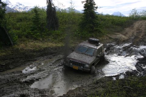 2003 Jeep TJ - A brown Jeep Cherokee XJ navigates a muddy trail, with a spare tire mounted on its roof.