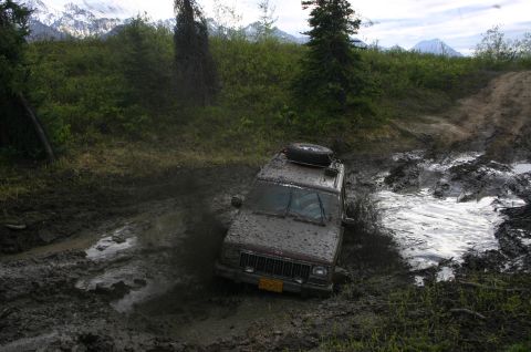 2003 Jeep TJ - A Jeep Cherokee XJ is stuck in deep mud on an off-road trail, with mountains visible in the distance.