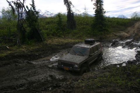 2003 Jeep TJ - A Jeep Cherokee navigates a challenging muddy trail with snow-capped mountains visible in the distance.