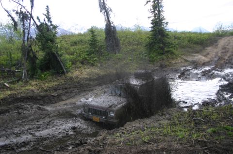 2003 Jeep TJ - A Jeep Cherokee XJ navigates a challenging muddy trail, kicking up a large spray of mud.