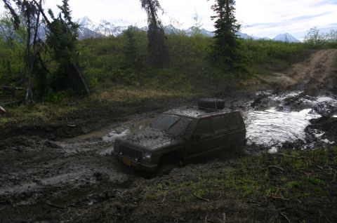 2003 Jeep TJ - A dark Jeep Cherokee XJ is stuck deep in mud on a trail with mountains in the background.