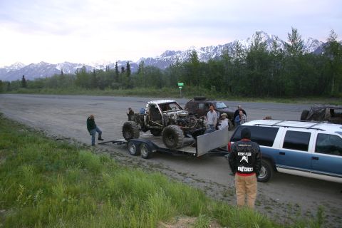 2003 Jeep TJ - Several vehicles are gathered near a trailer carrying a heavily modified off-road vehicle, with a mountainous landscape in the distance.