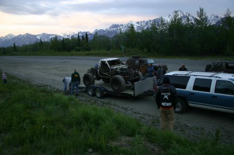 2003 Jeep TJ - Several people gather around a custom off-road vehicle being transported on a trailer, with a blue SUV and mountains visible in the distanc…