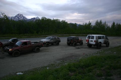 2003 Jeep TJ - A group of off-road vehicles are parked on a dirt road, with a backdrop of trees and snow-capped mountains under a cloudy sky.