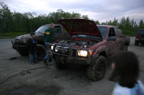 2003 Jeep TJ - Two mud-covered trucks are parked off-road, one with its hood open and headlights illuminated.