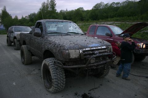 2003 Jeep TJ - Several mud-covered trucks are parked on a dirt road, with one having its hood open.
