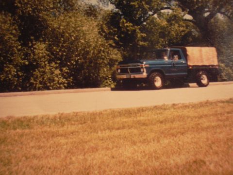 1977 Ford F-150 - A blue 1977 Ford F-150 flatbed truck is seen driving on a road with trees in the background.