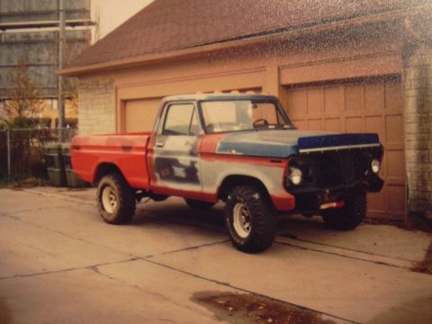 1977 Ford F-150 - A red and blue pickup truck with primer patches on the doors is parked in front of a garage.