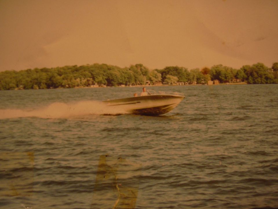 Speed boat cutting through water with a person at the helm. Trees and buildings line the distant shore