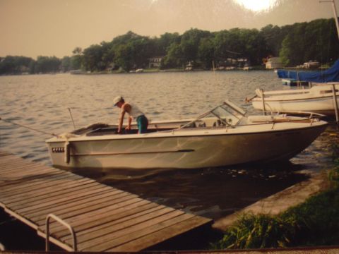 1977 Ford F-150 - A person is tending to a boat docked at a wooden pier on a calm lake, with another boat visible in the background.