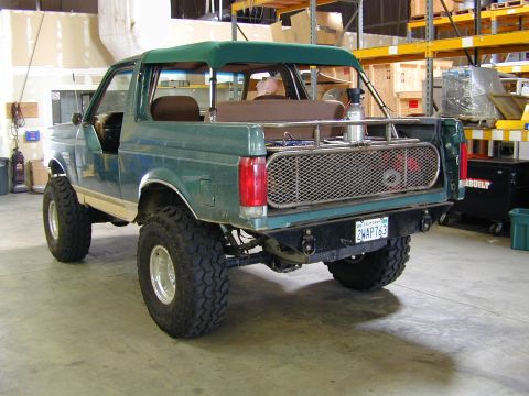 1989 Ford Bronco - Modified 1989 Ford Bronco seen from the rear in a workshop setting. It has large off-road tires and a custom bumper.