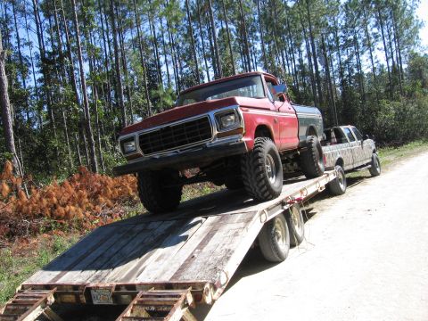 1974 Ford F-100 - A red pickup truck is being transported on a trailer, accompanied by another camouflaged truck, with a wooded area in the background.