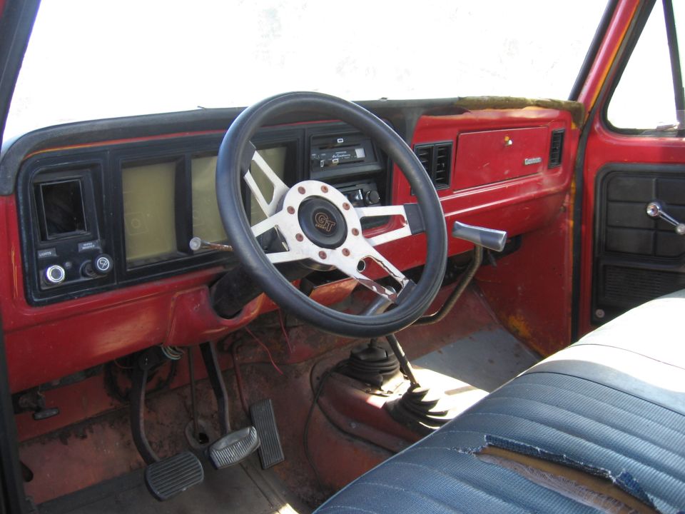 Interior of a truck cab, showing the dashboard, steering wheel, pedals, and part of the seat. The steering wheel has a GT logo