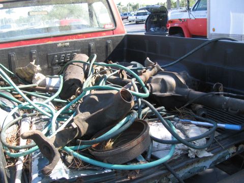1974 Ford F-100 - A red pickup truck bed is loaded with various rusty automotive parts and green hoses, with other vehicles in the background.