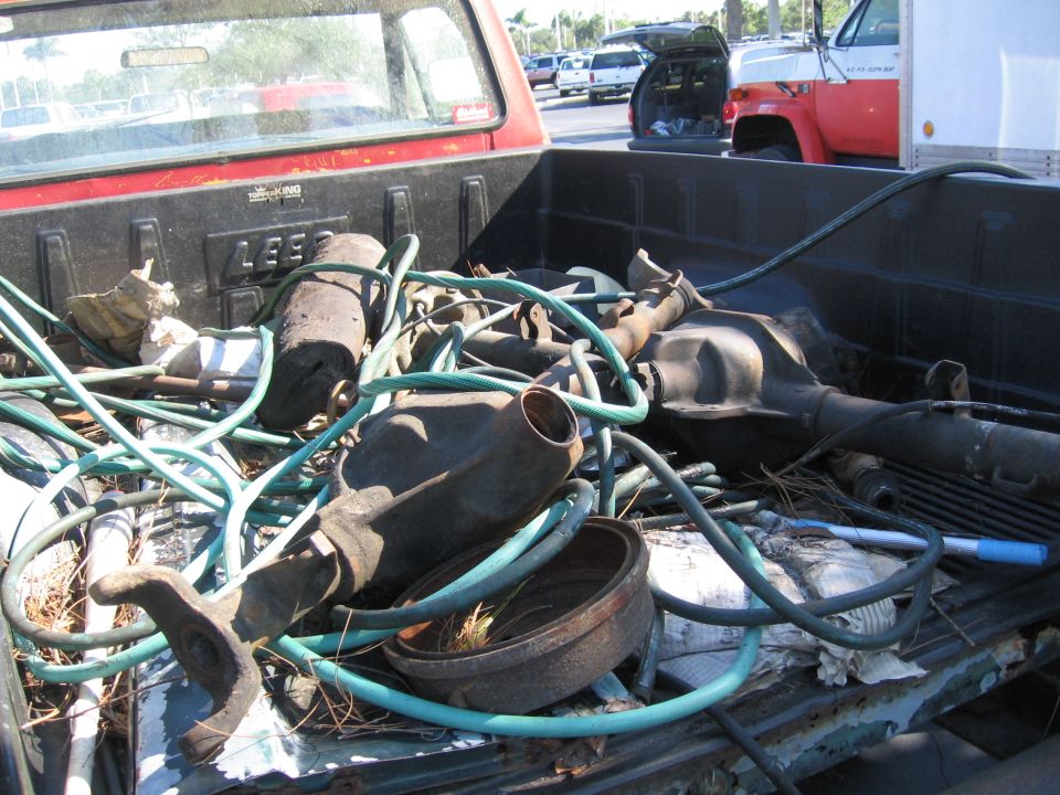 Red pickup truck bed filled with rusty auto parts and hoses. Another truck is visible in the background
