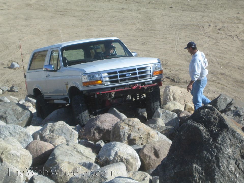 Silver Ford Bronco with lifted suspension navigating a rocky terrain. A man stands nearby