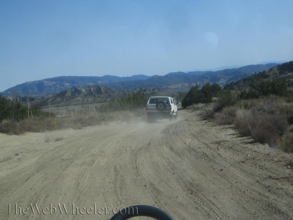 White 1987 Suzuki Samurai driving on a dusty dirt road with mountains in the background