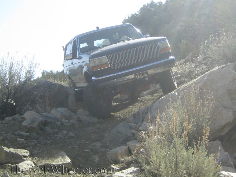 Dark Ford Bronco climbing rocks on a trail. Has a chrome grille and a sticker on the rear bumper