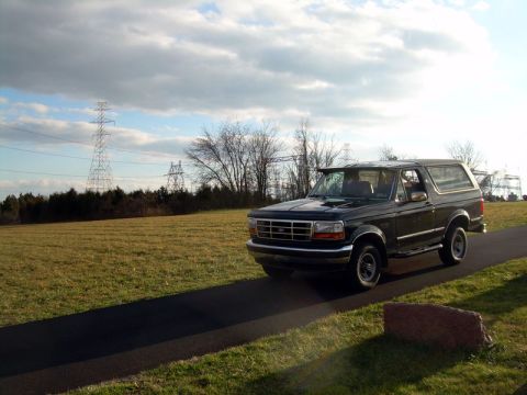 1992 Ford Bronco - A black 1992 Ford Bronco is shown driving on a paved path with a field and power lines in the background.