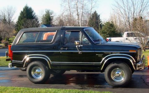 1984 Ford Bronco - A black Ford Bronco SUV is parked on a wet driveway with a white pickup truck in the background.