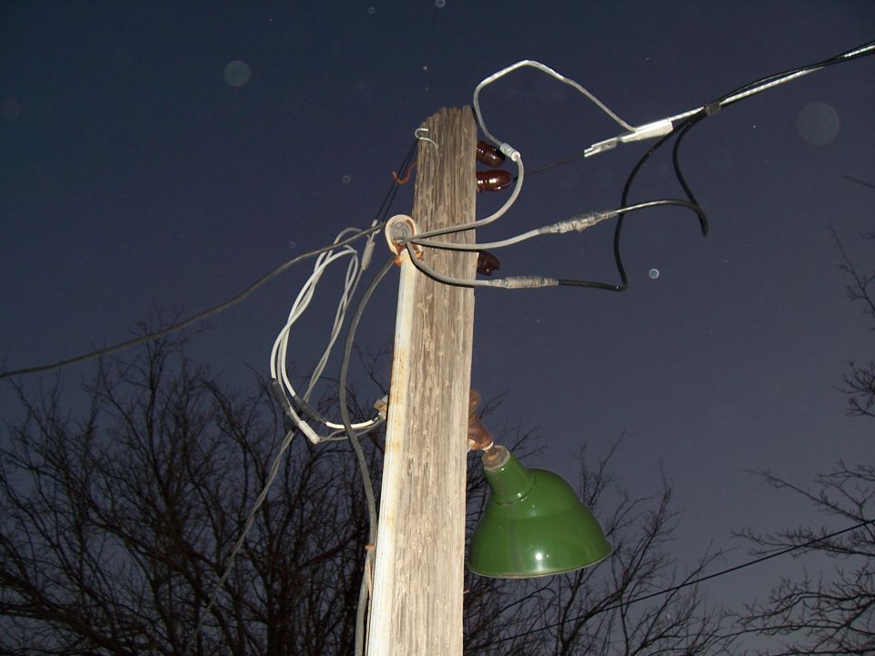 Electrical pole with tangled wires and a green lamp against a dark sky