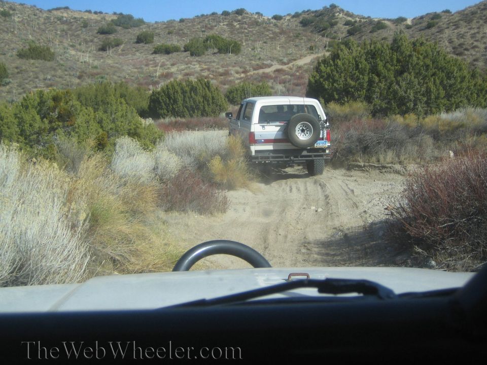 White SUV with spare tire on back driving up a dusty dirt road in a desert landscape. Another vehicle is behind it