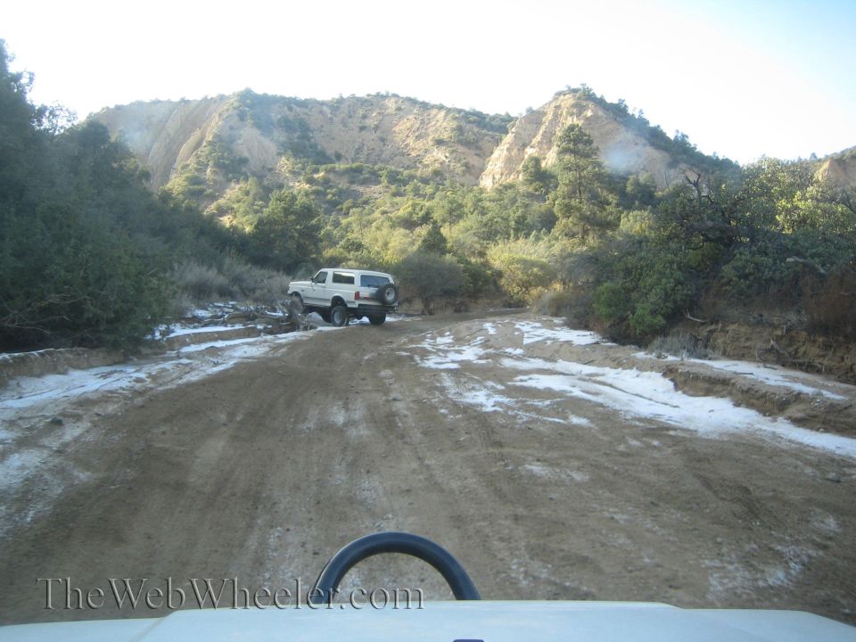 White SUV parked on a dirt road with snow patches, surrounded by trees and hills
